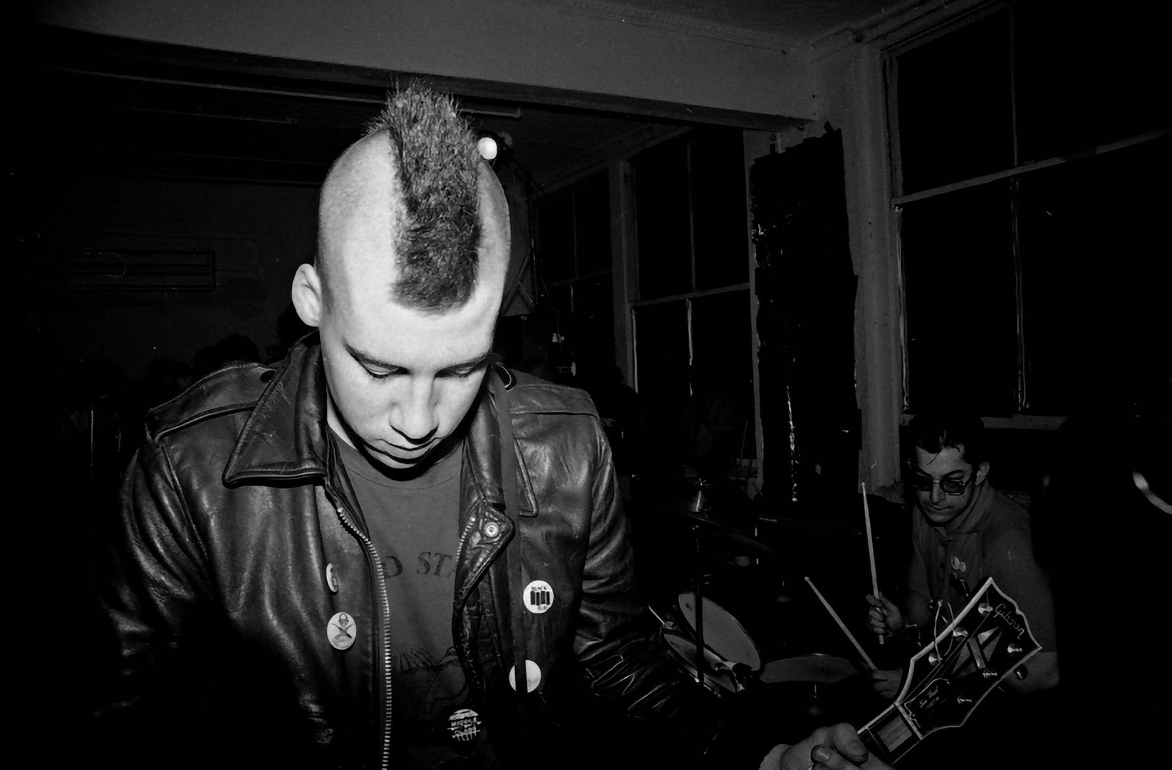 Schwarzweißaufnahme eines Punkmusikers mit Gitarre und Schlagzeuger im Hintergrund